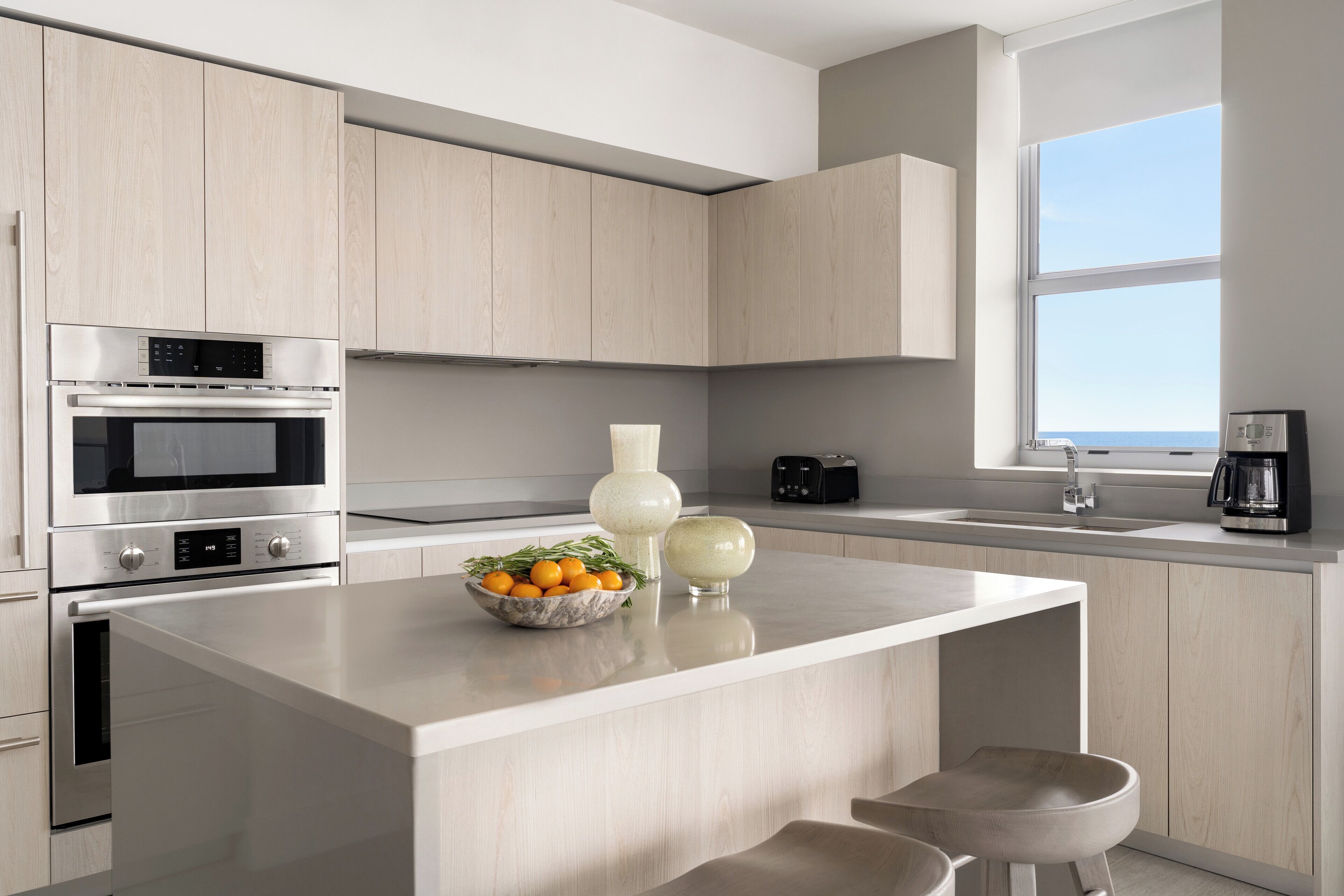A modern kitchen with light wood cabinets, stainless steel appliances, a central island with seating, and a view of the sea through a large window.