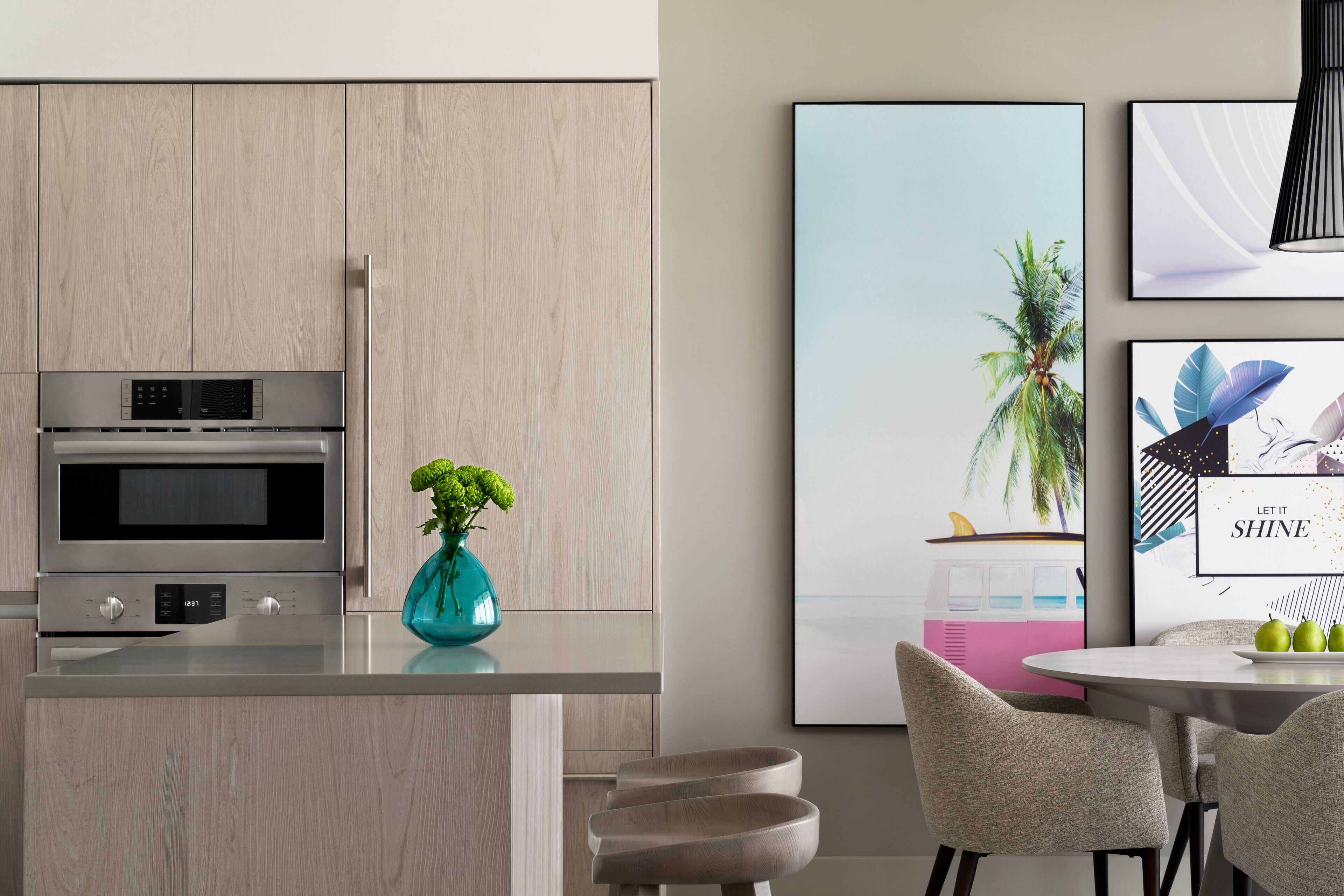 A modern kitchen with a built-in oven and microwave, a teal vase with green plants, and a dining area with a round table and chairs.