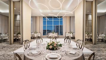 A banquet hall with round tables set for a formal event, large windows with a city view, and modern lighting fixtures.