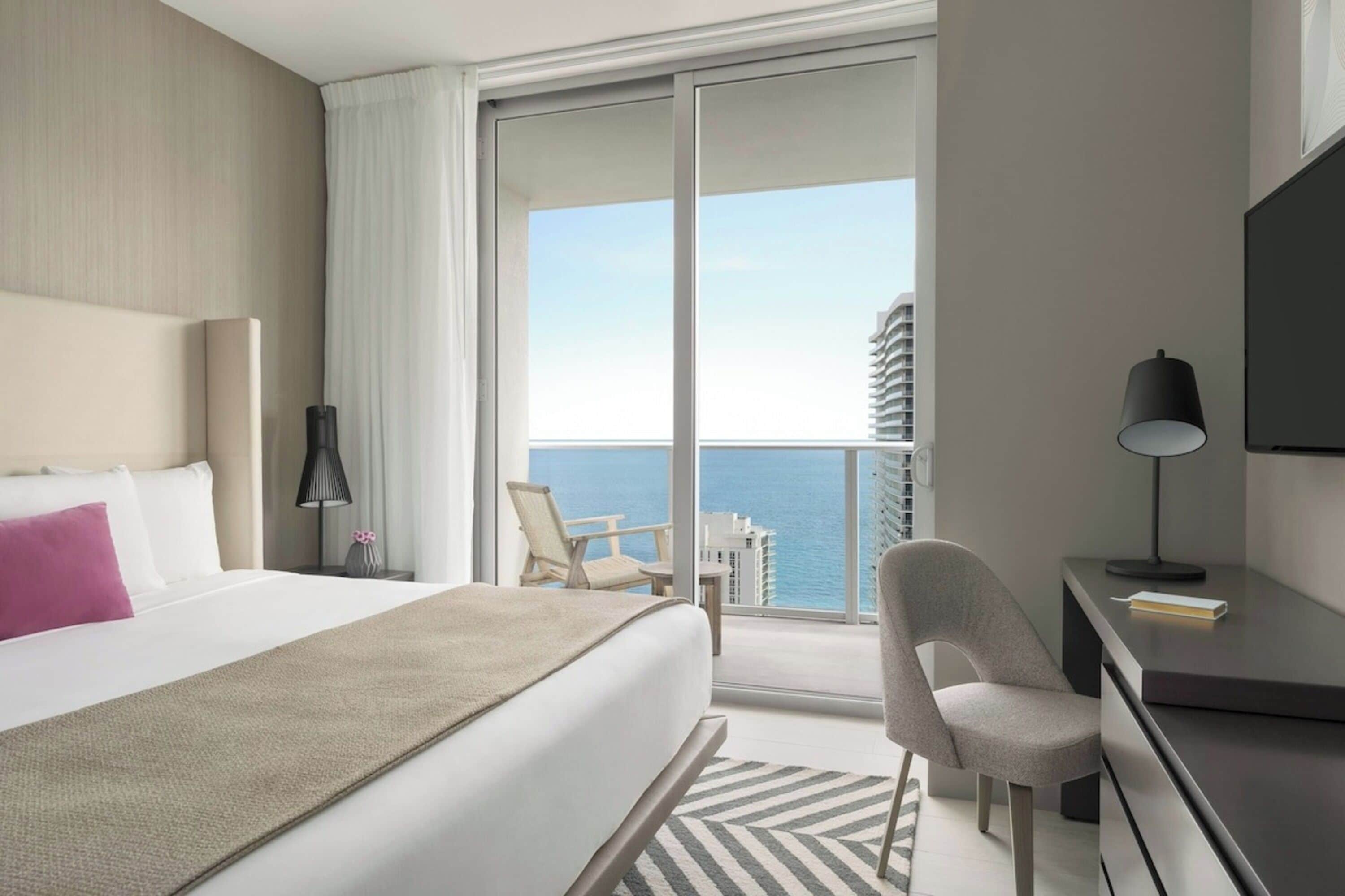 A hotel room with a bed, a desk, a chair, a lamp, and a view of the ocean.