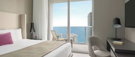 A hotel room with a bed, a desk, a chair, a lamp, and a view of the ocean.