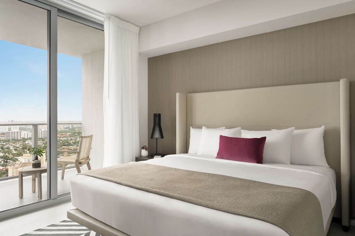 Une chambre d’hôtel moderne avec un grand lit, une table de chevet, une lampe, une chaise et une vue sur la ville par une grande fenêtre.