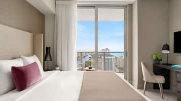 Habitación de hotel moderna con una cama grande, un escritorio, una silla, un televisor y vistas a la ciudad y al agua.