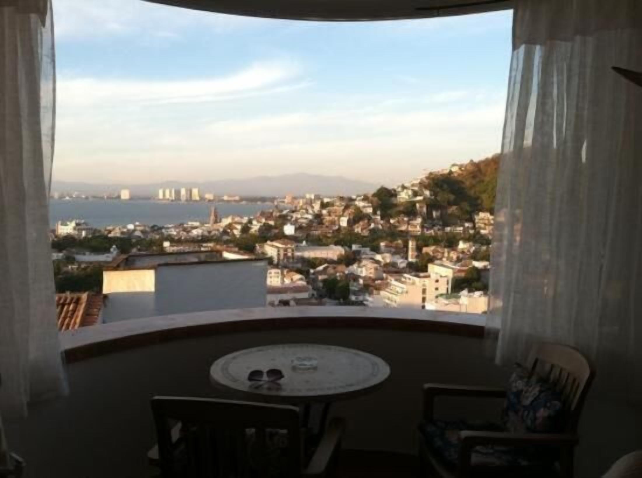 A balcony with a table and chairs, overlooking a coastal town with buildings and mountains in the distance.