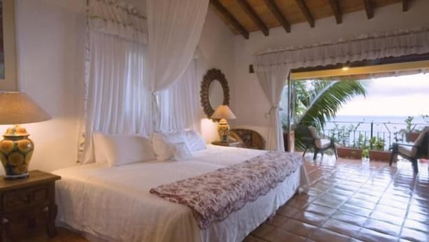 Un dormitorio con cama, cortinas, una mesita de noche, lámpara, espejo y vista al mar.