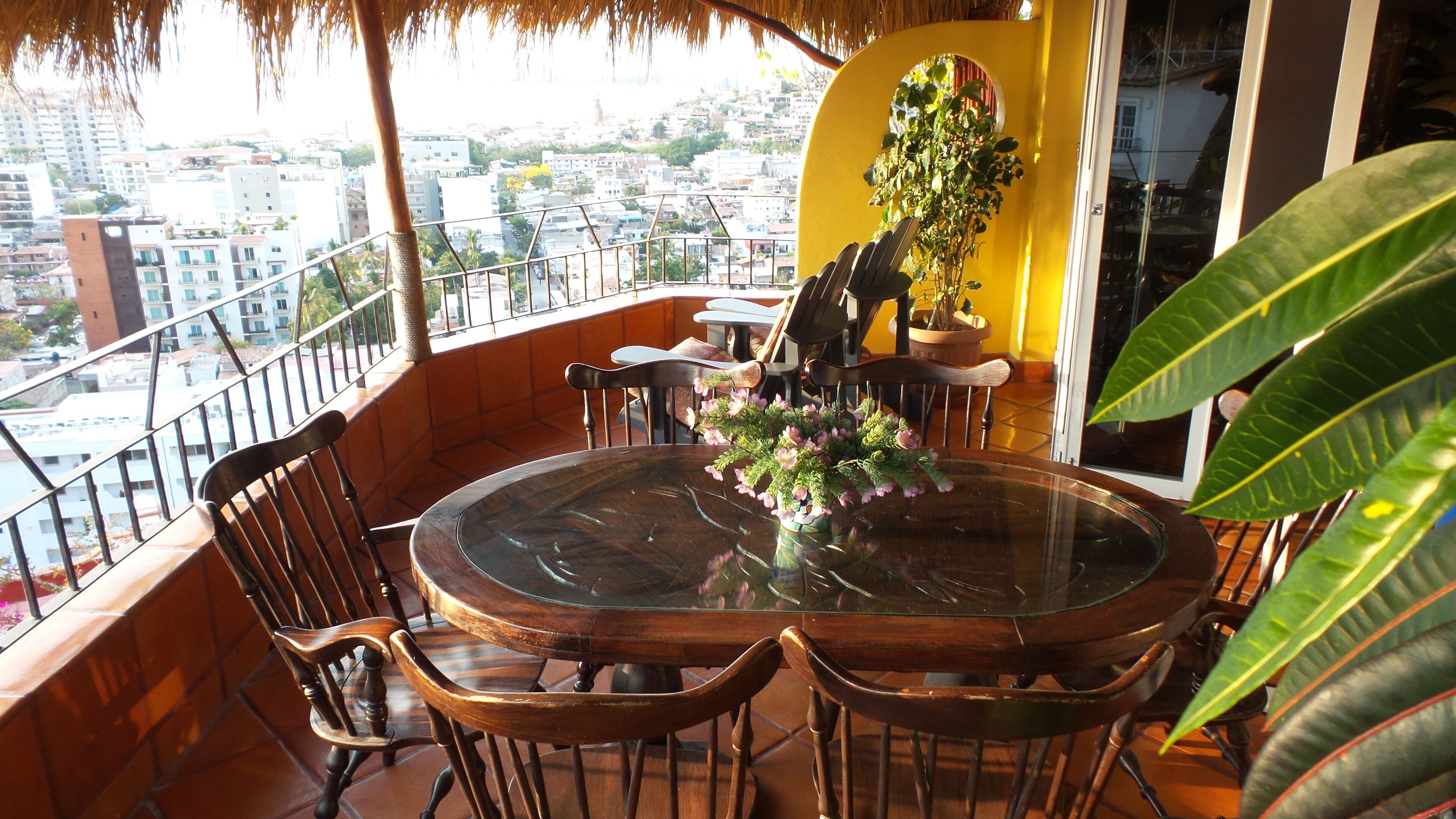 Une table ronde en verre, entourée de chaises, sur un balcon donnant sur une vue panoramique de la ville.