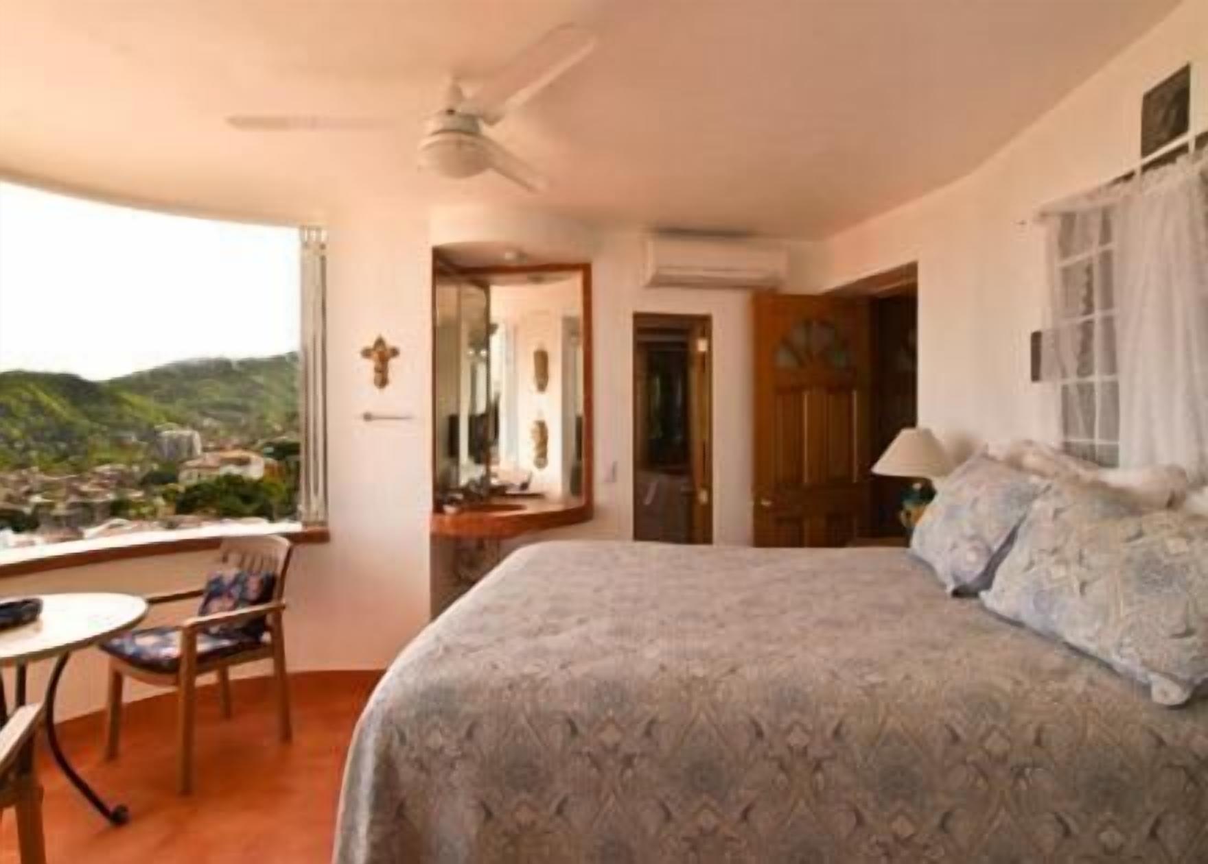 A bedroom with a large bed, a chair, a table, and a view of a cityscape through a window.