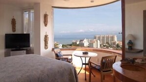 Un dormitorio con cama, televisión y balcón con vista a la ciudad y al océano.