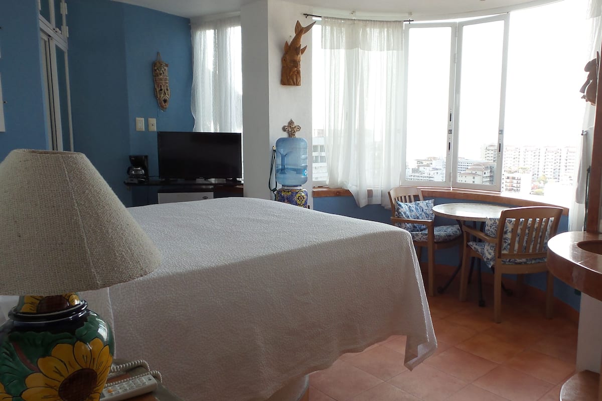 A hotel room with a bed, a desk, a chair, a table, a water dispenser, a television, and a large window with curtains.