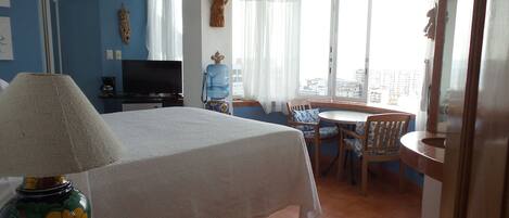 A hotel room with a bed, a desk, a chair, a table, a water dispenser, a television, and a large window with curtains.