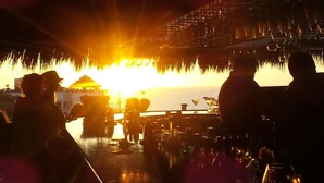 Un bar con vistas a la puesta de sol sobre el agua, siluetas de personas y una estructura de edificio tradicional.