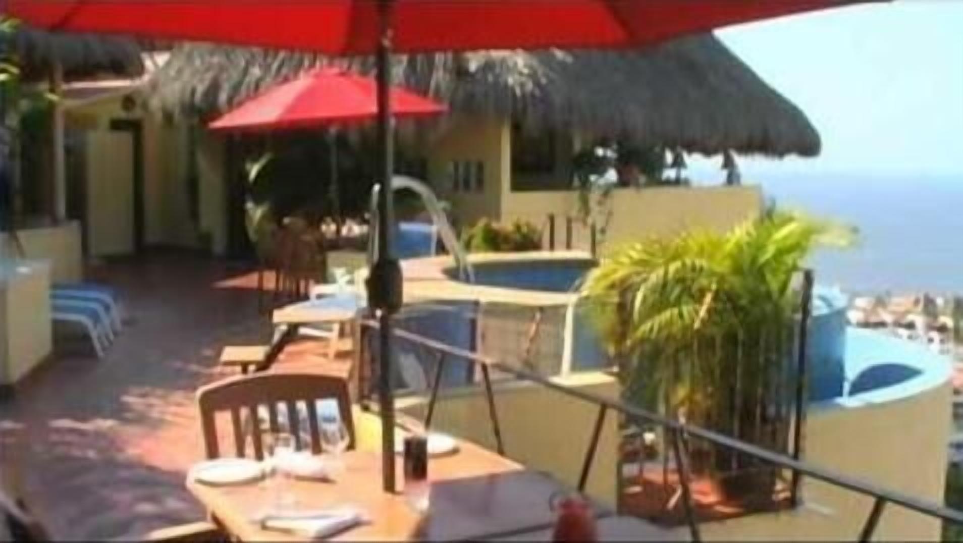 An outdoor dining area with tables and chairs, a red umbrella, and a pool in the background.
