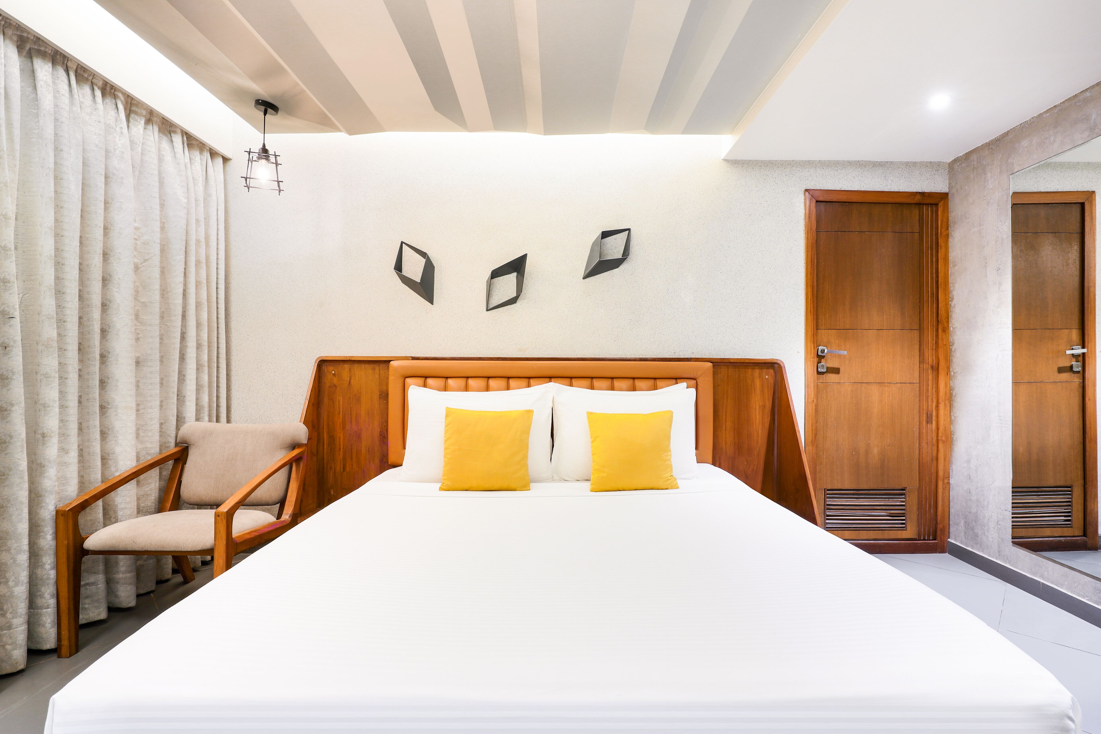 Habitación de hotel con una cama grande, cabecera de madera, dos almohadas amarillas, una silla de madera, una puerta de madera y una lámpara.