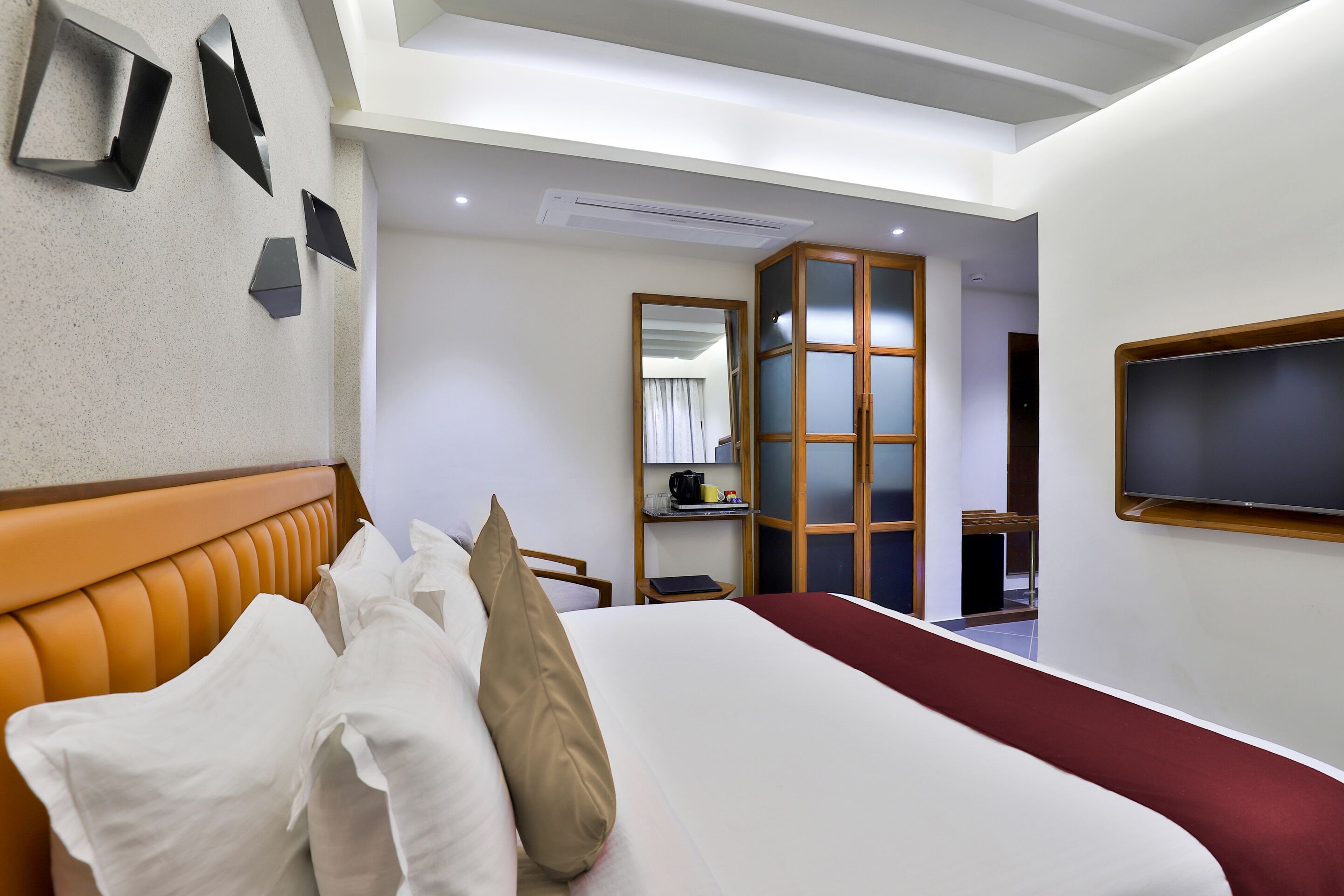 A hotel room with a bed, pillows, a TV, a mirror, and a shelf with items.