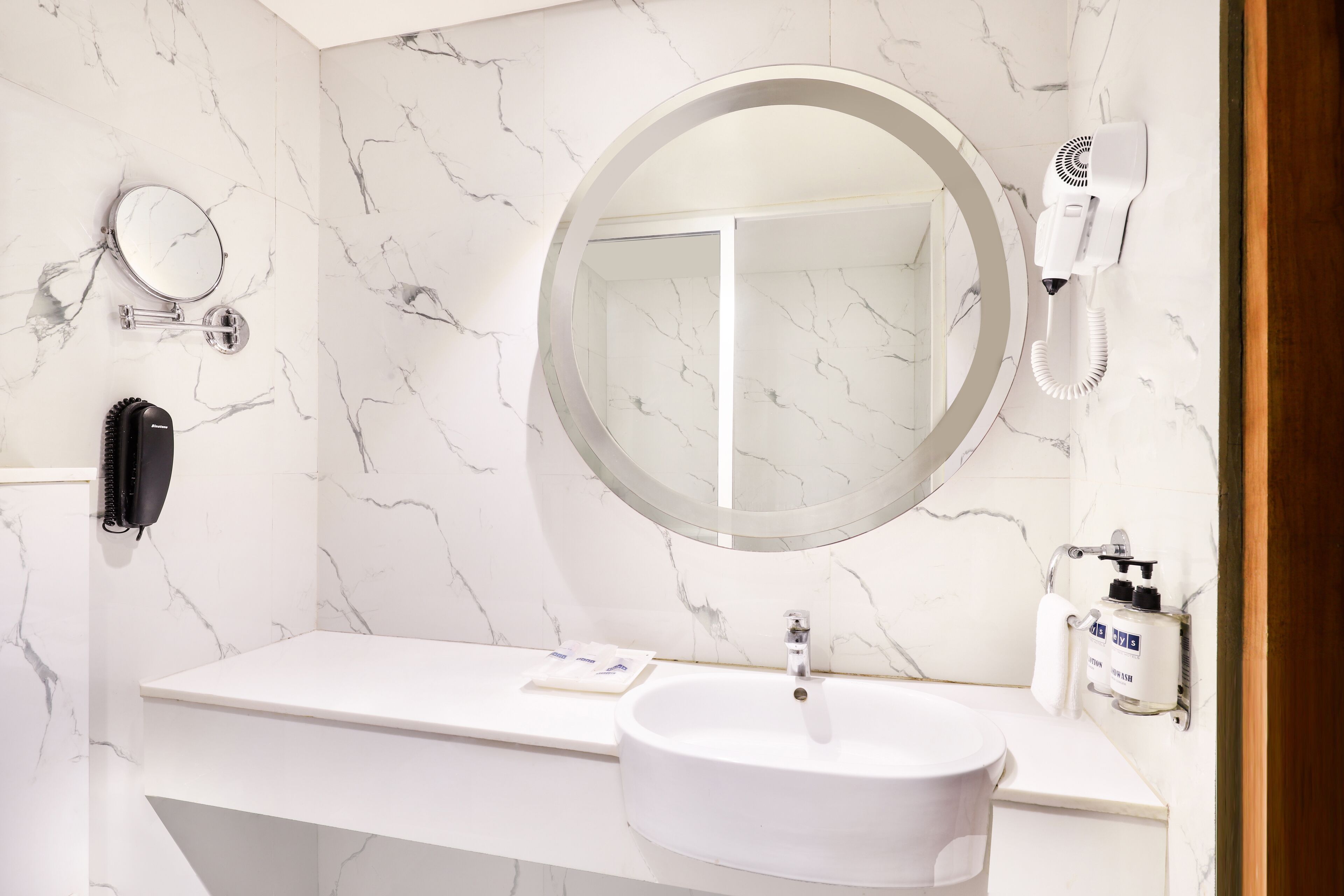 A bathroom with a white marble wall, a round mirror, a white sink, a hairdryer, and a telephone.