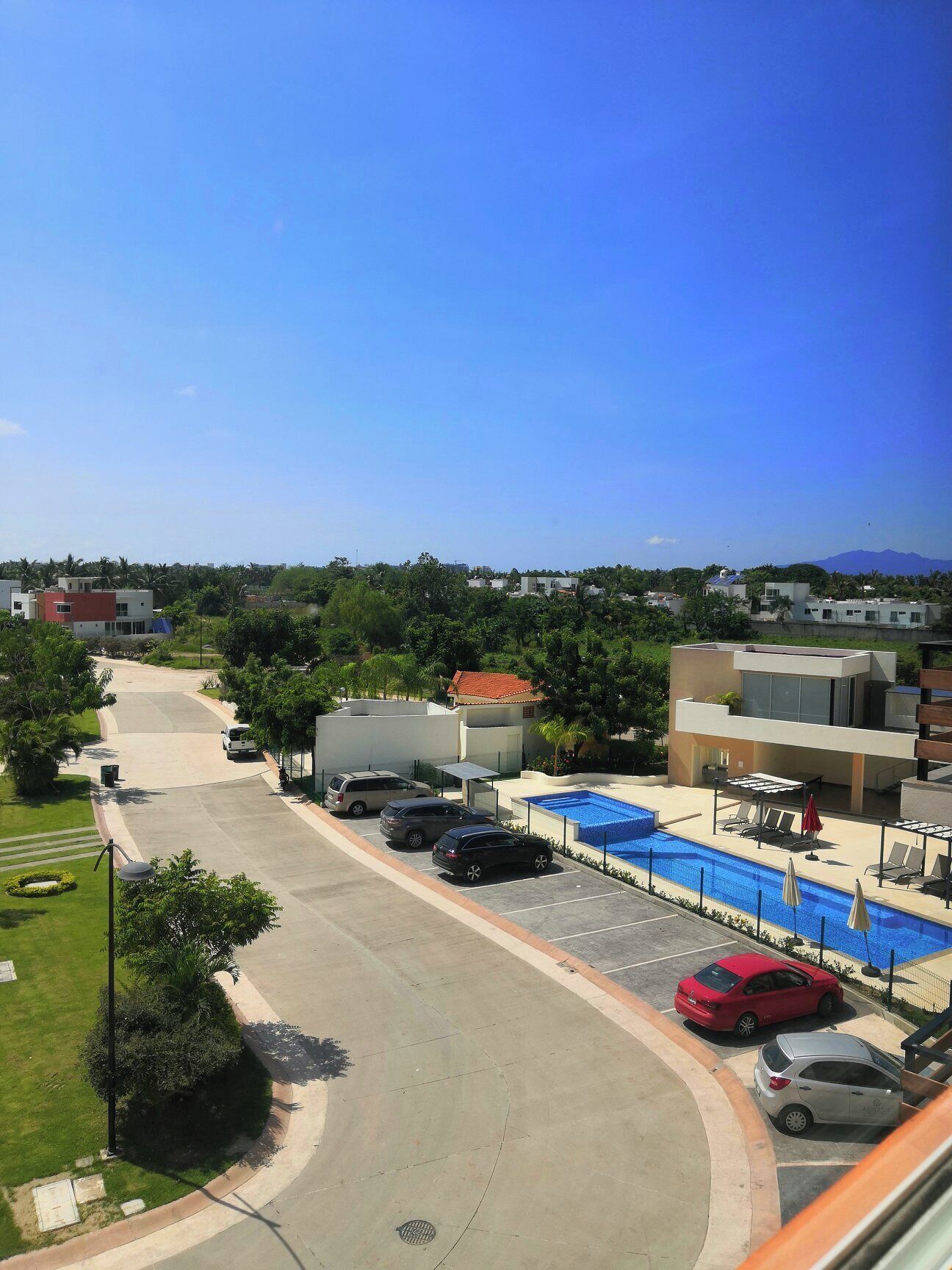 A parking area with a swimming pool, a covered seating area, and a view of a residential area with houses and greenery.