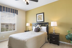 A bedroom with a bed, two bedside tables with lamps, a ceiling fan, a window with blinds, and a framed picture on the wall.