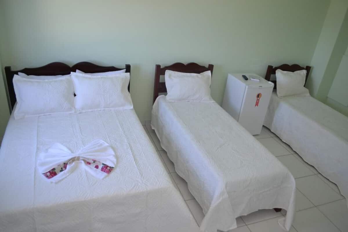 Ein Hotelzimmer mit drei Einzelbetten, jedes mit weißer Bettdecke und Kissen. Ein kleiner Kühlschrank steht auf dem Boden zwischen den Betten.