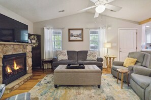 A living room with a fireplace, a sofa, an ottoman, a television, and a ceiling fan. - Welcome to “Relax Inn” Retreat at Lake Cumberland – A Perfect Cabin Escape (Burnside)
