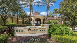 A landscaped entrance with a sign for Windsor Hills Clubhouse, surrounded by palm trees and well-maintained hedges. - Summer Settings by VillaDirect (Kissimmee)