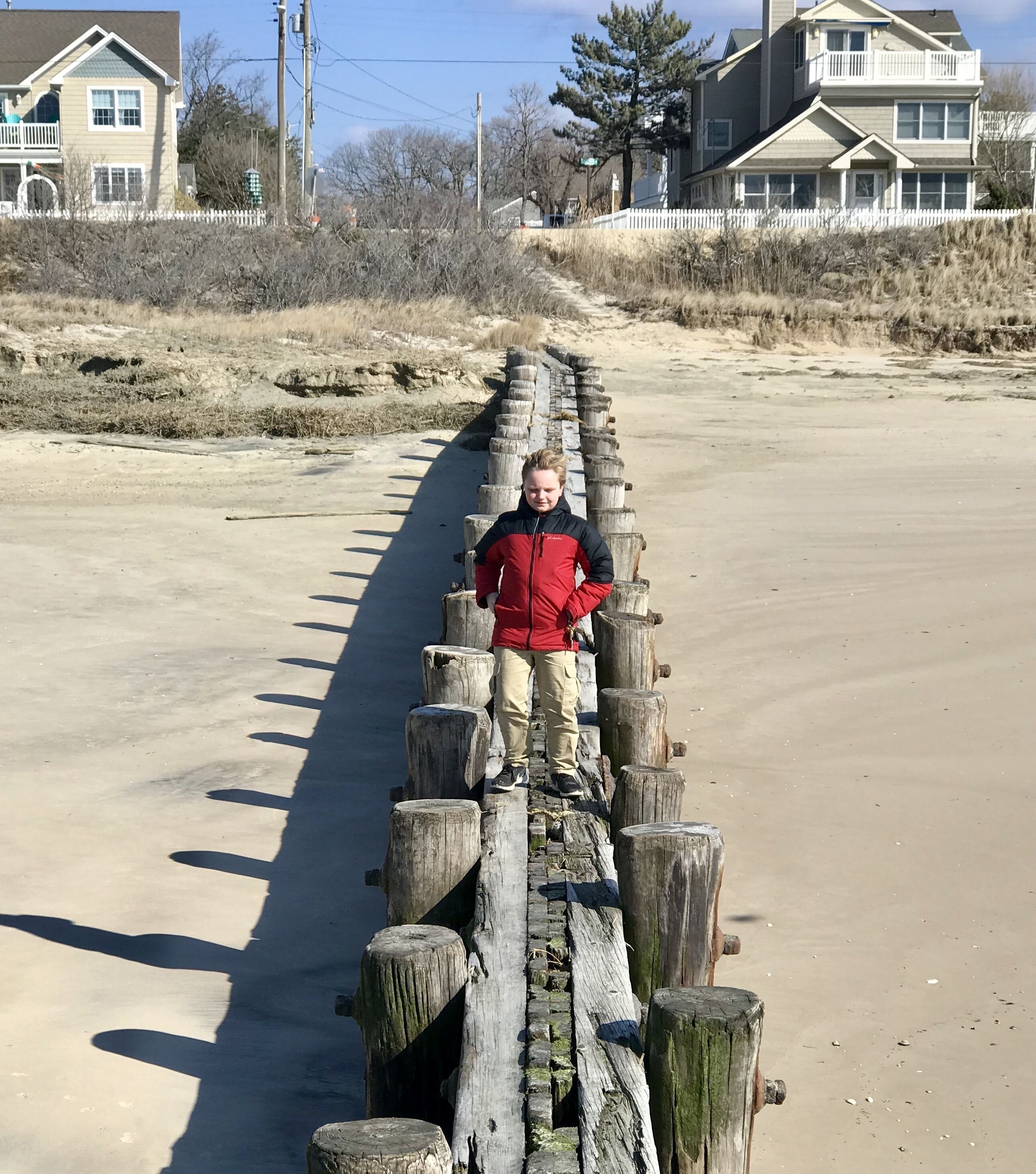 A person standing on a wooden boardwalk by a sandy beach with houses in the background.