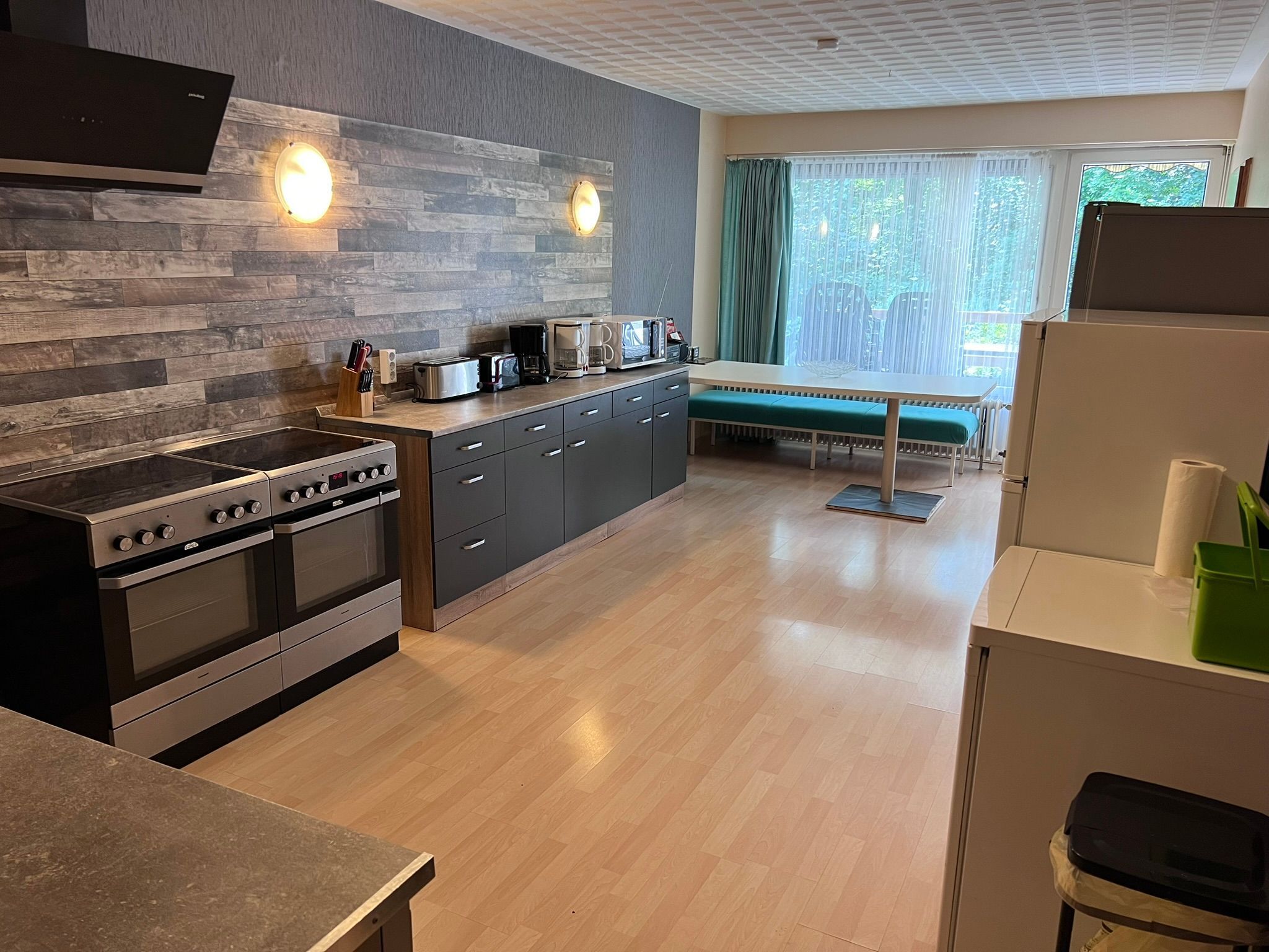 A modern kitchen with a built-in oven, a dining area with a bench, and a view of greenery outside.