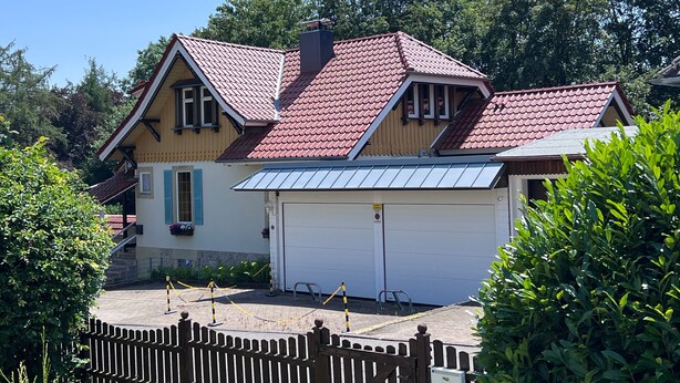 Une maison à deux étages avec un toit recouvert de tuiles rouges, un garage et une allée bordée d’une clôture.