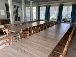 A long dining table with multiple chairs, large windows with curtains, and a view of greenery outside.