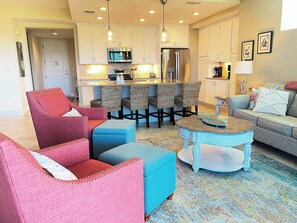 A modern living room with a kitchen in the background, featuring a dining area with wicker chairs, a round coffee table, and a sofa.