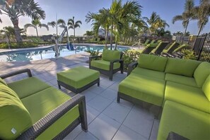 Zona junto a la piscina con sillones de descanso y otomanas, palmeras y cielo despejado.