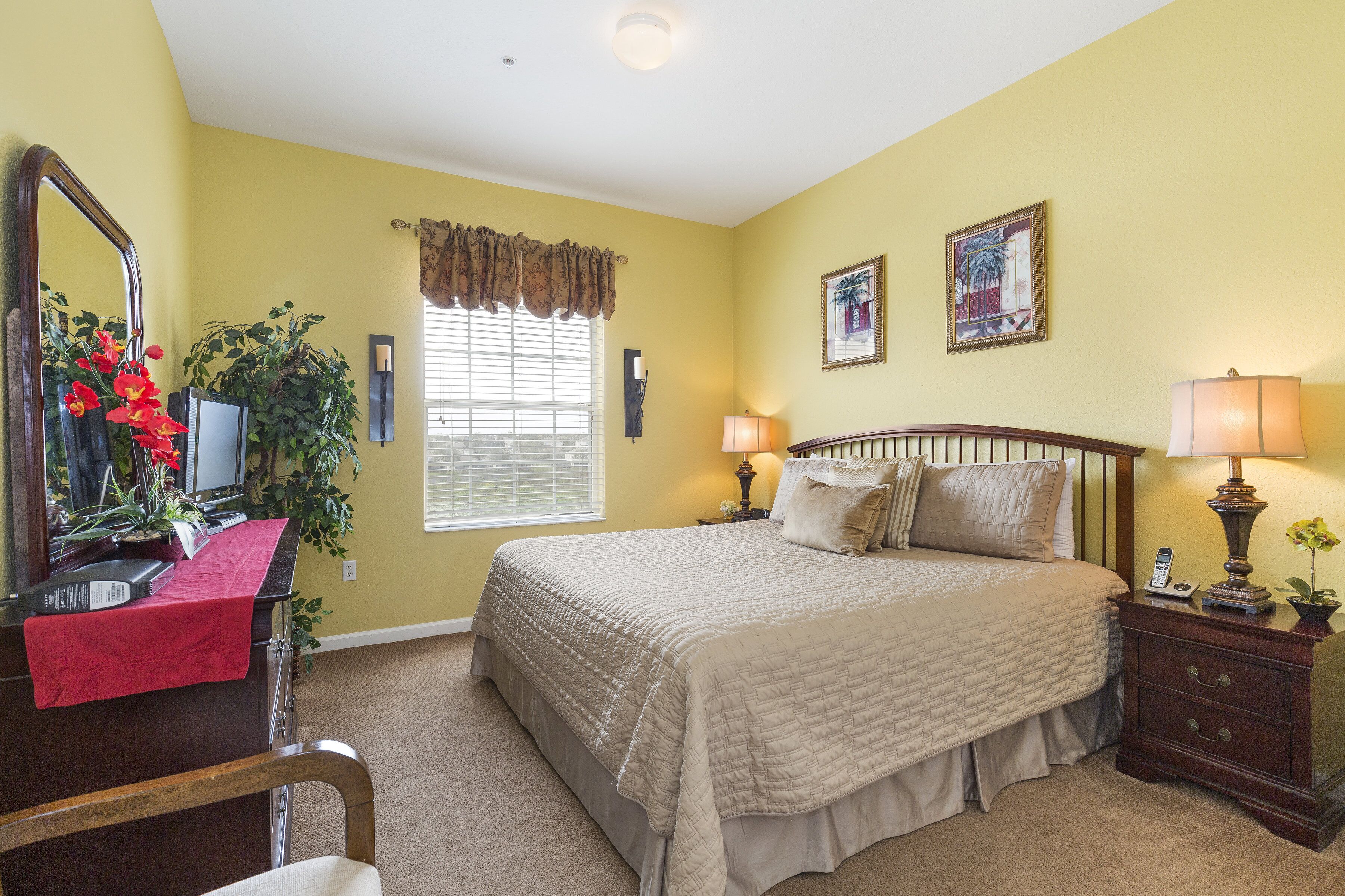 A bedroom with a bed, bedside tables, a mirror, a window with blinds, and a plant.