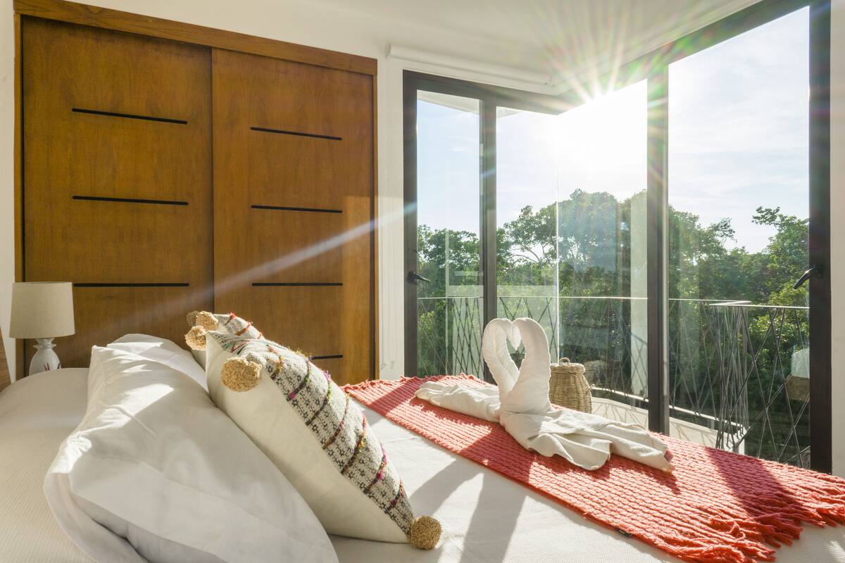 Habitación de hotel con un ventanal grande, una cama con ropa blanca, un armario de madera y una alfombra roja.