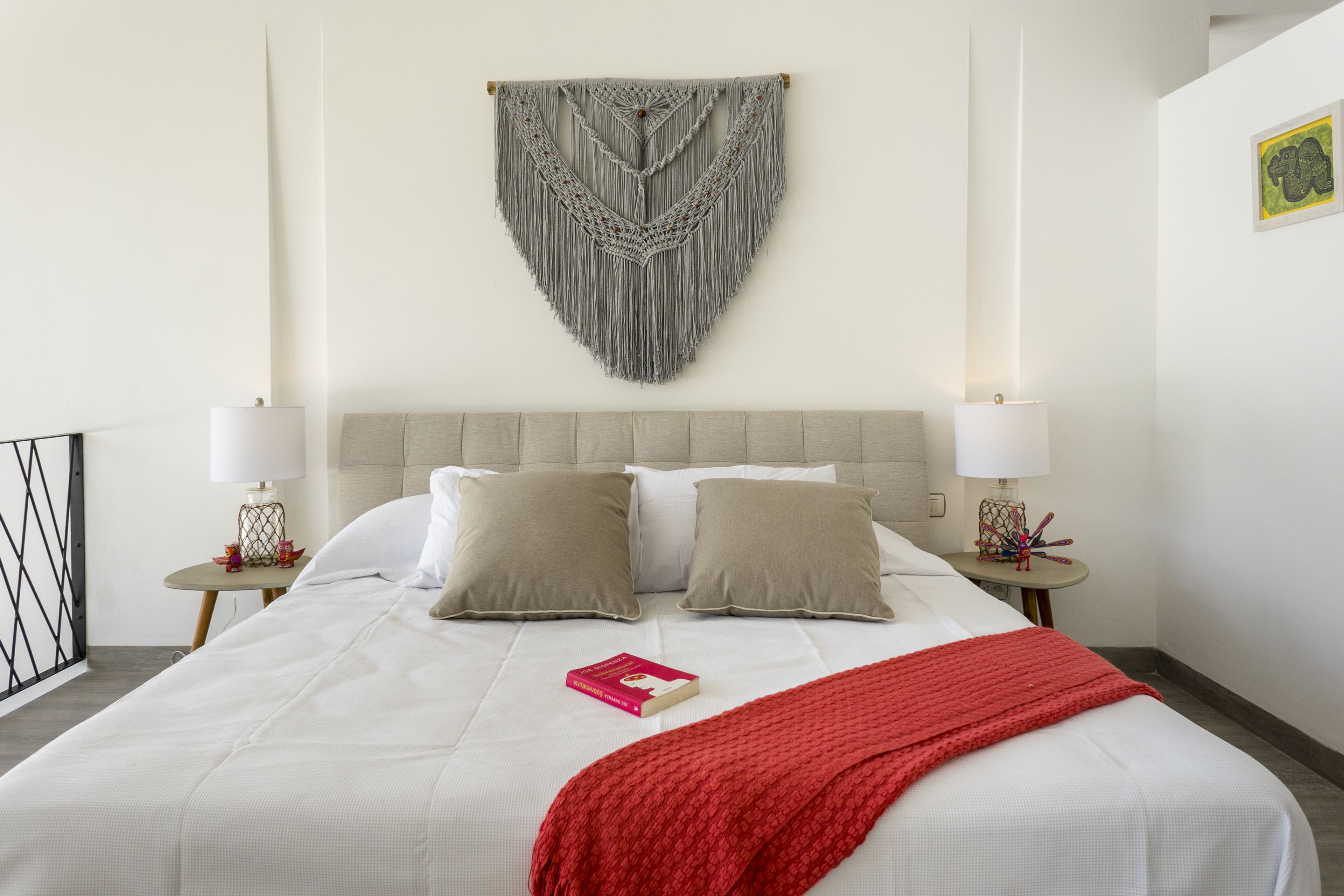 A neatly made bed with a red blanket, two pillows, and a macrame wall hanging above.