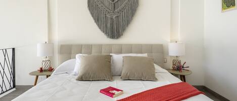 A neatly made bed with a red blanket, two pillows, and a macrame wall hanging above.