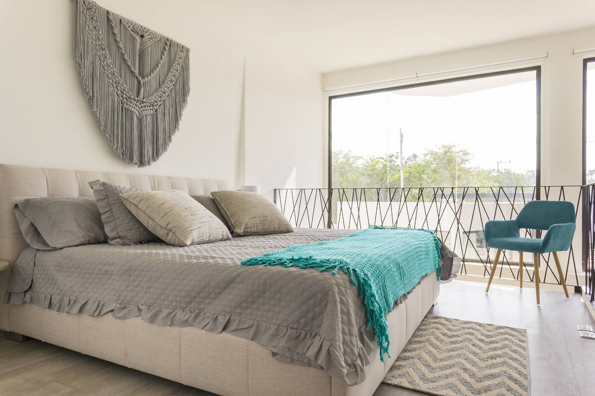 Un dormitorio con una cama, un tapiz de macramé, vistas a través de un ventanal grande y una silla color turquesa.