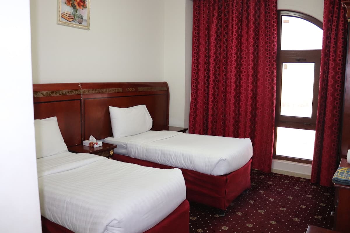 Une chambre d’hôtel avec deux lits simples, une tête de lit en bois, une petite table de chevet avec une boîte de mouchoirs et une fenêtre avec des rideaux rouges.