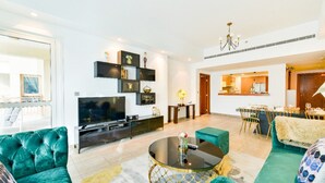 A modern living room with a large flat-screen TV, teal sofas, a coffee table, and a dining area with a chandelier.