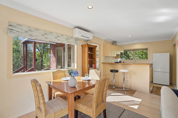 A dining area with a wooden table and wicker chairs, a kitchen with a white refrigerator, and a window with a view of trees.