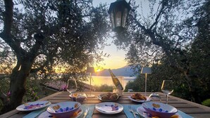 A wooden table set for a meal with plates, bowls, cutlery, and glasses, overlooking a scenic sunset and mountains. - Lake Garda luxury studio with pool (Torri del Benaco)