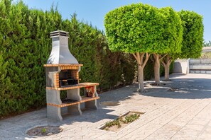 An outdoor grill with a chimney, surrounded by a paved patio, a neatly trimmed tree, and a hedge. - Fidalsa Grand White House (Mutxamel / Muchamiel)