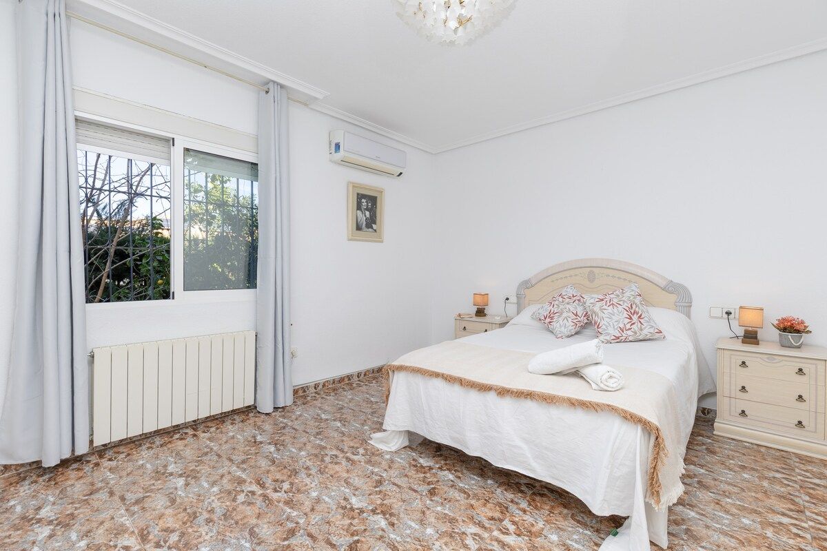 A bedroom with a bed, bedside tables, a radiator, a window with curtains, and a picture on the wall.
