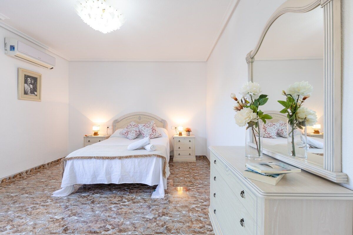 Ein Schlafzimmer mit einem Bett, Nachttischlampen, einem Schminktisch, einem Spiegel und einem gerahmten Bild an der Wand.