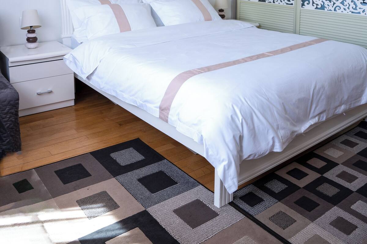 Un lit bien fait, avec une tête de lit blanche, une table de chevet avec une lampe et un tapis à motifs.