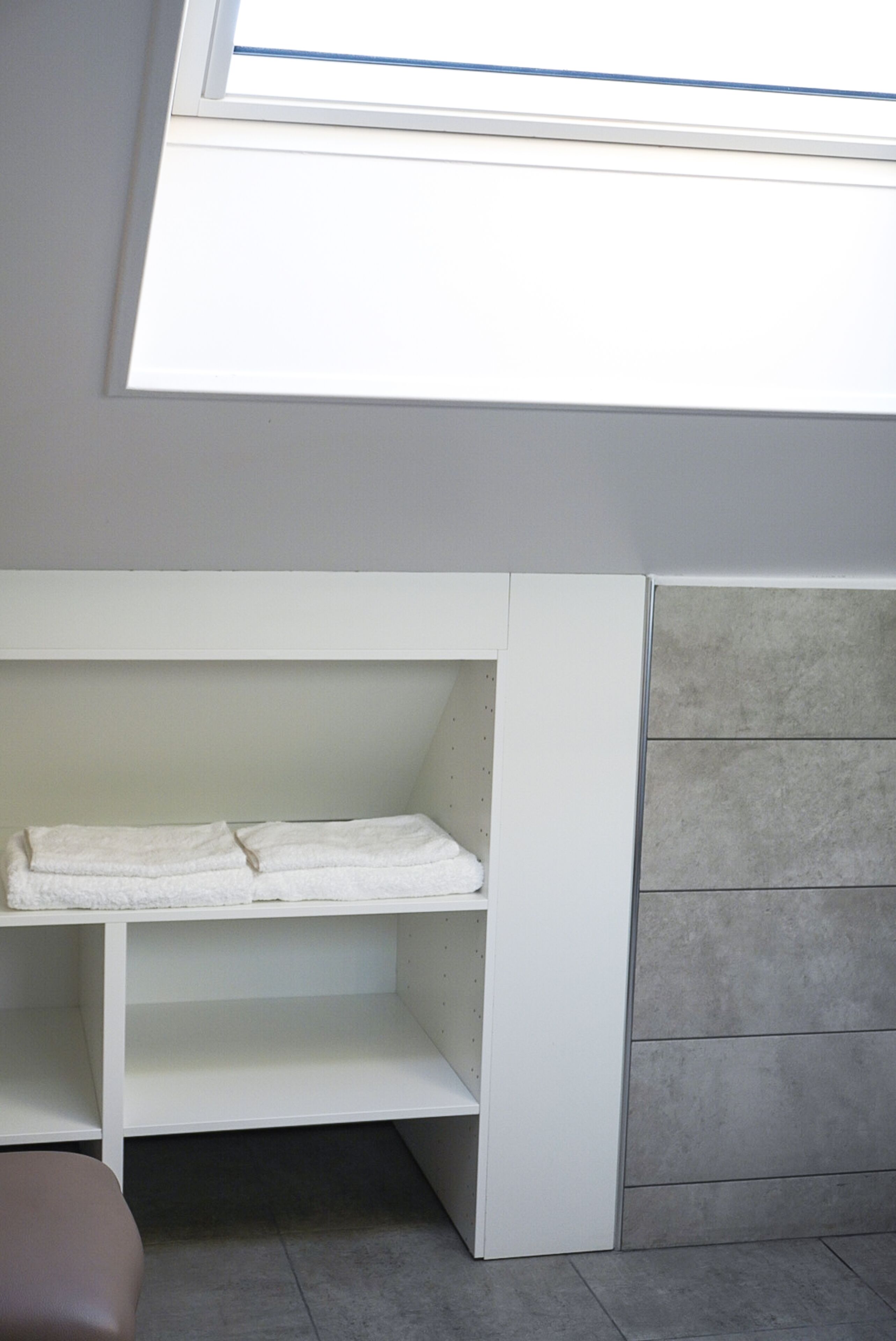 Una habitación con tragaluz, un banco empotrado blanco con estantes y paredes de baldosas grises.