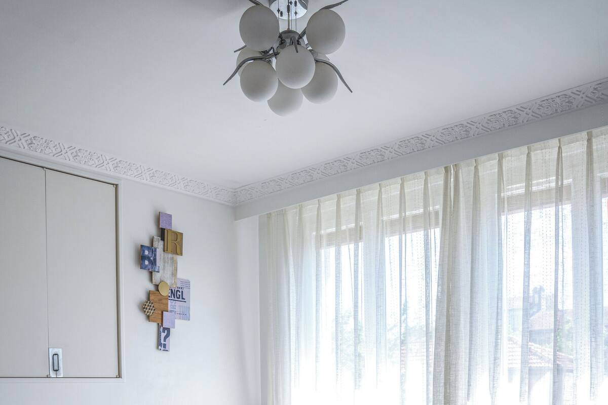 Une chambre à coucher bien rangée, avec un lit, un ventilateur de plafond et une fenêtre ornée de rideaux légers.