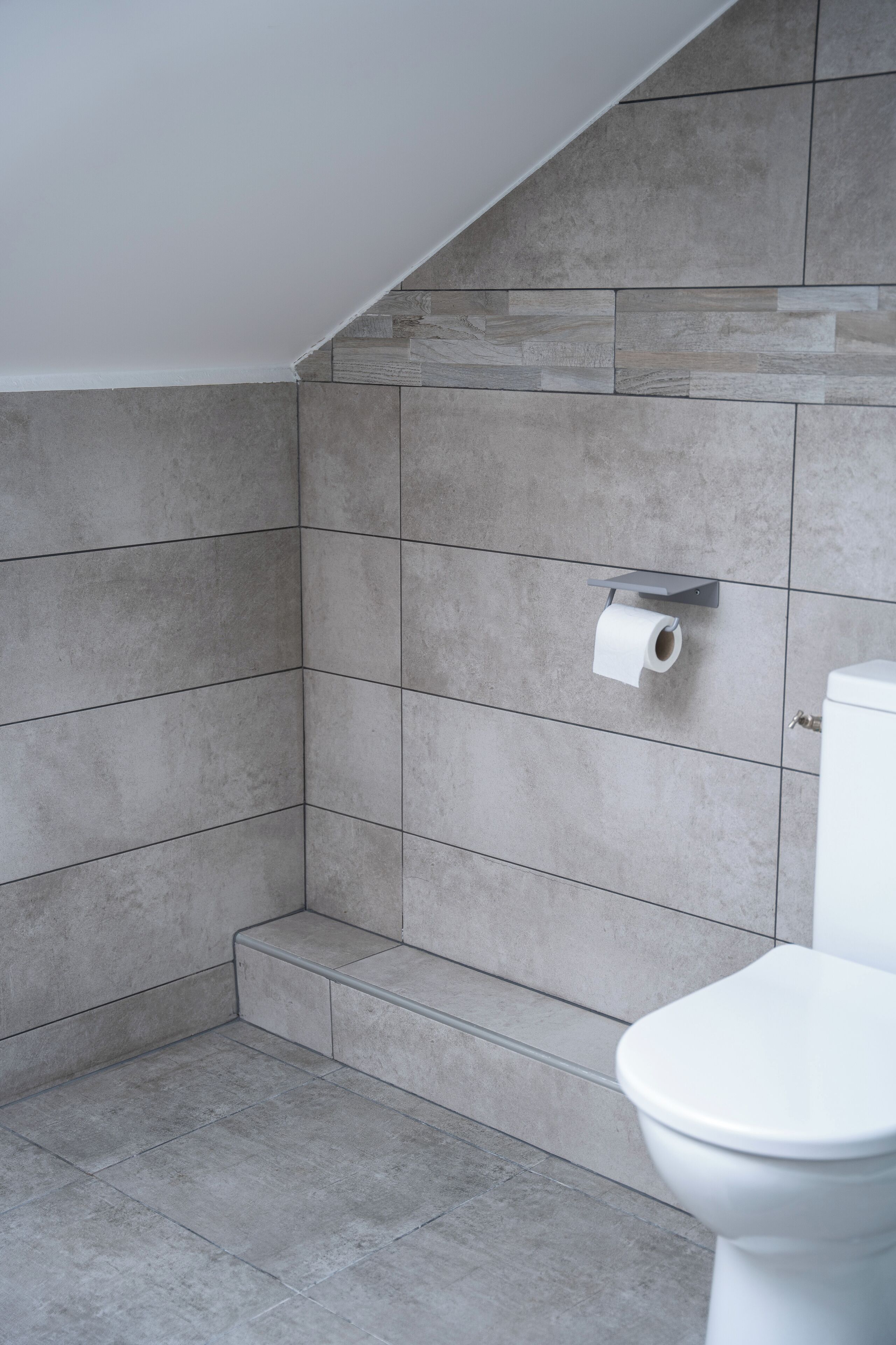 A bathroom with grey tiles, a toilet, and a toilet paper holder on a shelf.