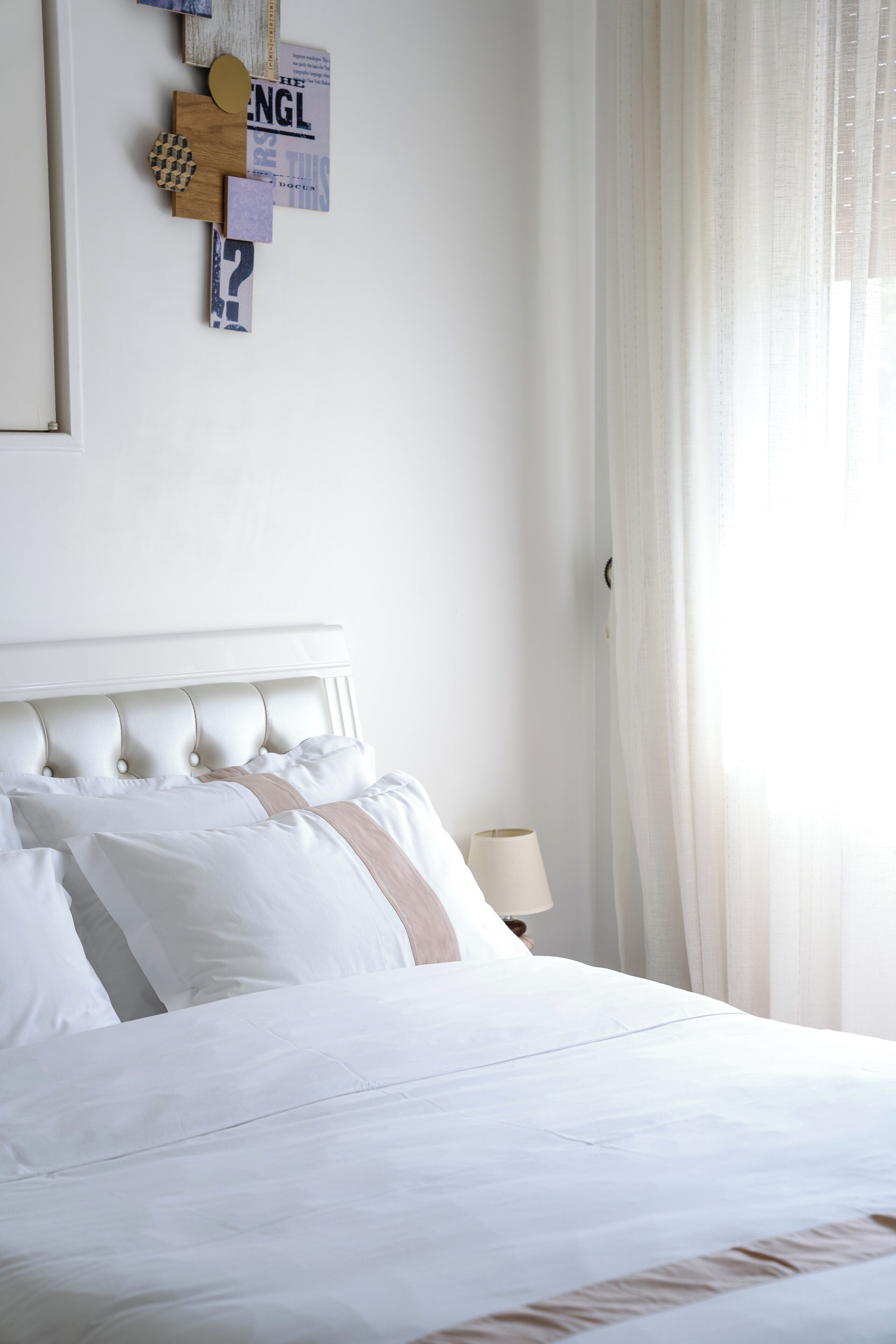 A neatly made bed with white and beige bedding, a wooden headboard, and a wall-mounted abstract art piece.