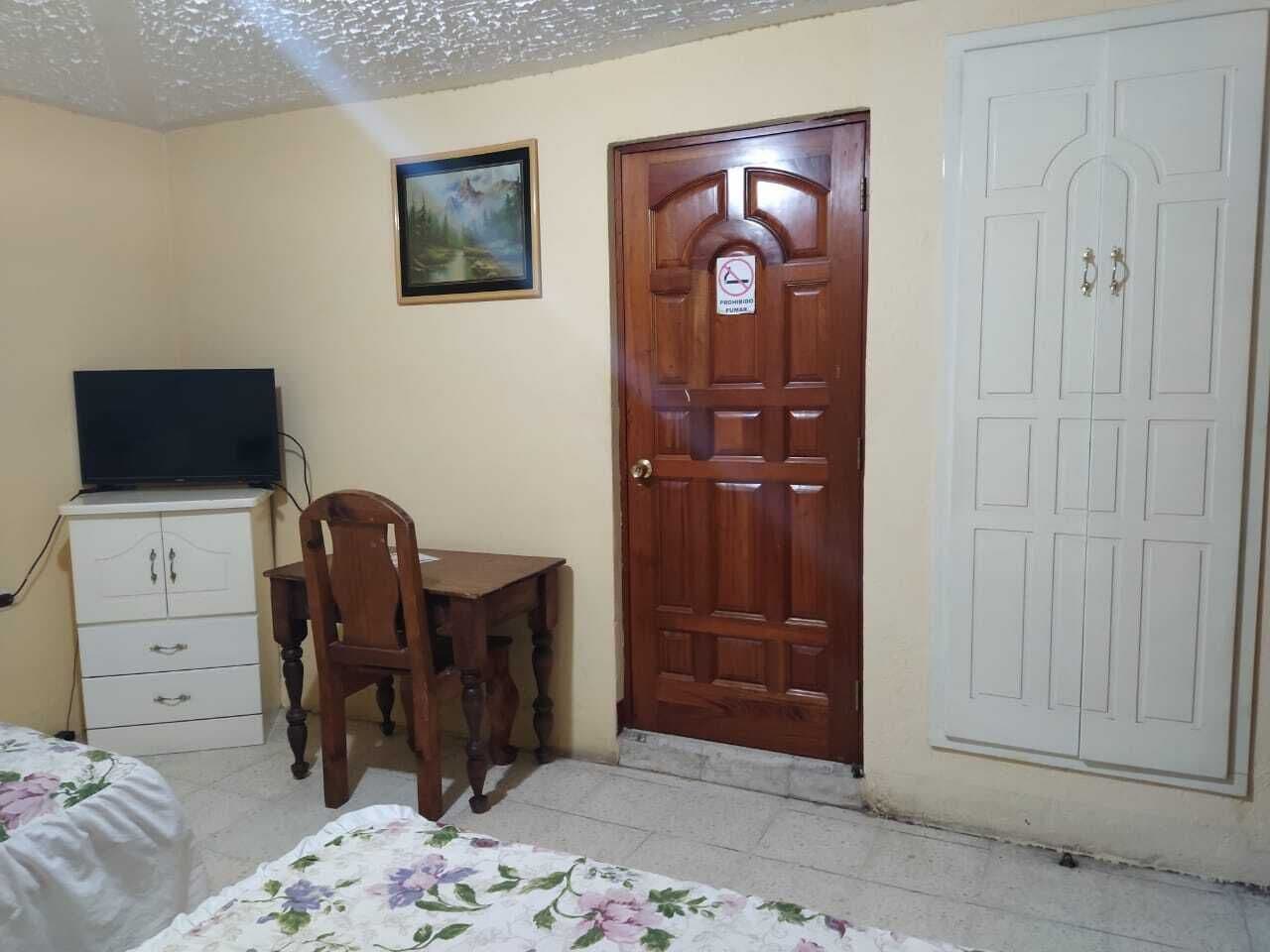 A bedroom with a bed, a wooden desk, a chair, a television on a cabinet, and two doors.