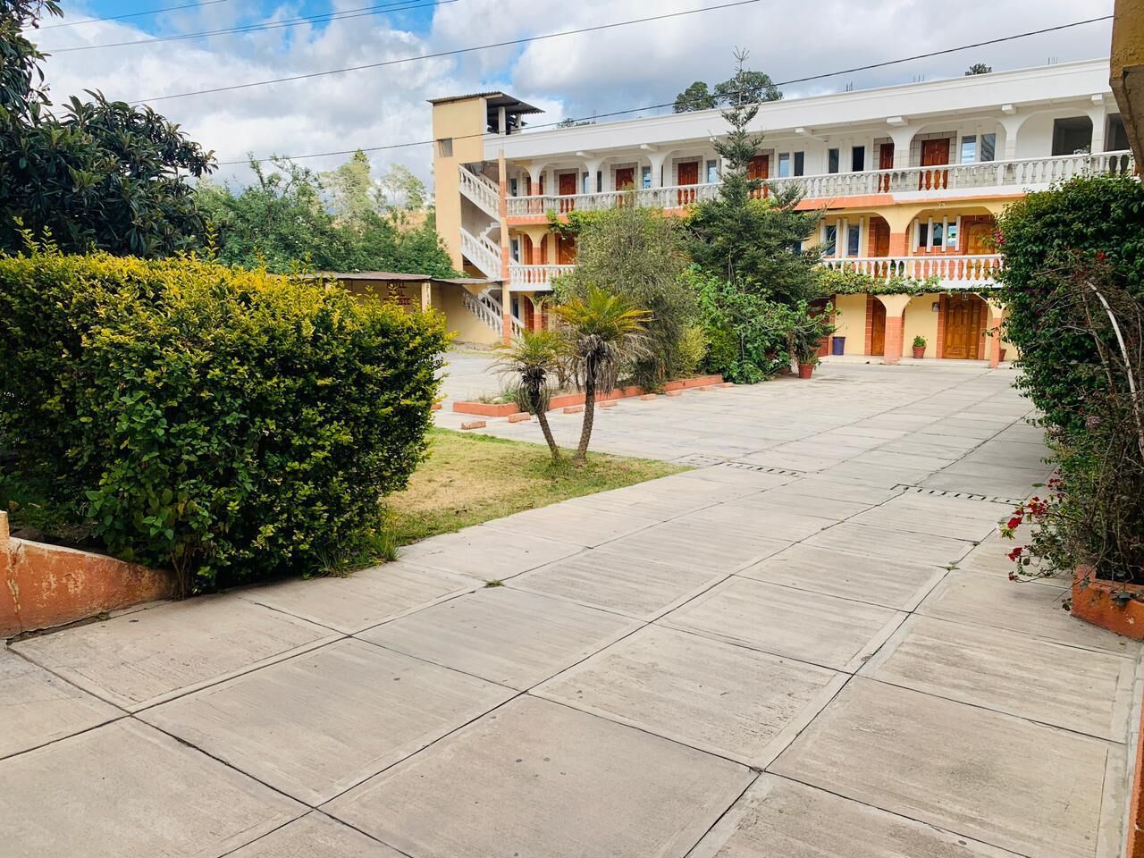 A courtyard with a paved walkway, surrounded by greenery and a multi-story building with balconies.