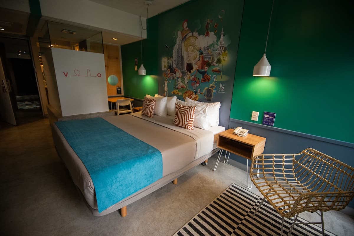 Una habitación de hotel moderna con una cama grande, un escritorio y una silla. Hay una pared verde con un mural y una pizarra con la palabra “Velo”.